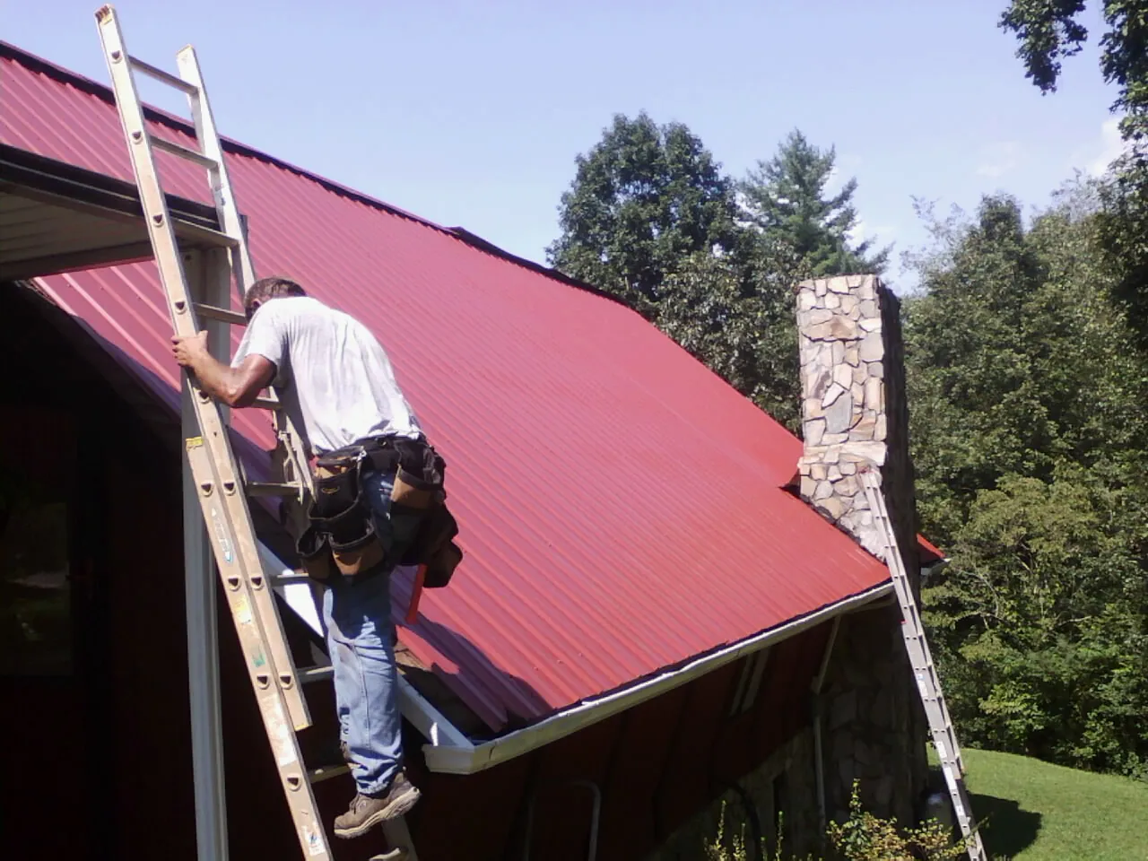 Roofer on a ladder beside a red metal roof and stone chimney — Soffit & Fascia Repair by Roofing Brothers in Shelter Island